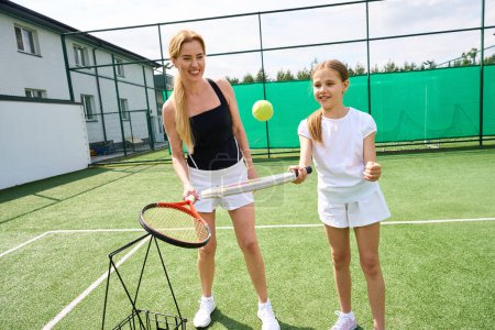 Foto de Alegre madre enseña a su hija a jugar al tenis, la chica golpea la pelota de tenis con una raqueta - Imagen libre de derechos