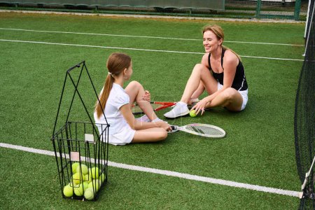Foto de Chica feliz con su madre están descansando en la hierba en la cancha de tenis, tienen raquetas de tenis y canasta con pelotas - Imagen libre de derechos