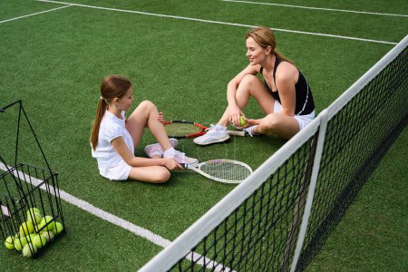 Foto de Mujer y su hija están descansando en la hierba en la cancha de tenis, tienen raquetas de tenis en sus manos - Imagen libre de derechos