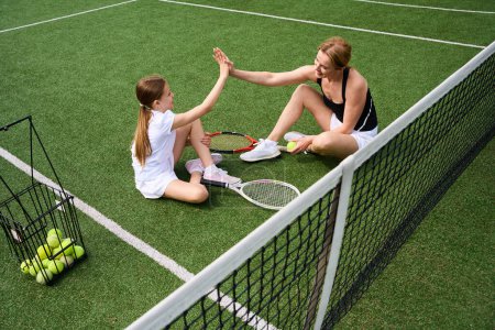 Foto de Adolescente y su madre se comunican en la hierba en la cancha de tenis, tienen raquetas de tenis y canasta de bolas - Imagen libre de derechos