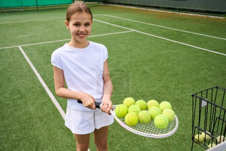 Photo for Teenage girl holding a lot of yellow balls on a tennis racket, green grass on the court - Royalty Free Image