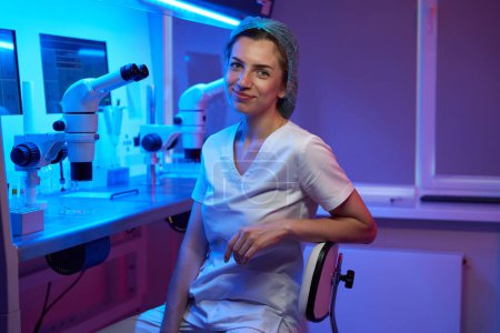 Foto de Un joven embriólogo con uniforme médico se sienta en el lugar de trabajo, sobre la mesa de un potente microscopio - Imagen libre de derechos