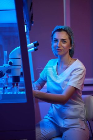 Téléchargez les photos : Jolie femme en uniforme médical assise sur le lieu de travail, sur le bureau d'un puissant microscope - en image libre de droit