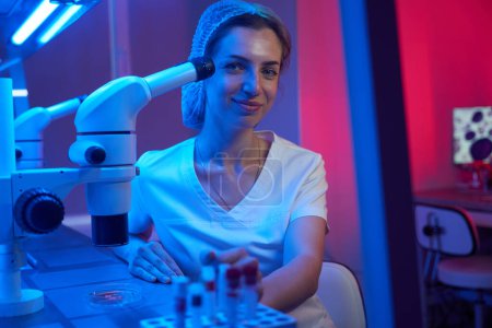 Foto de Un joven asistente de laboratorio se sienta en el lugar de trabajo en el laboratorio, sobre la mesa de un potente microscopio - Imagen libre de derechos