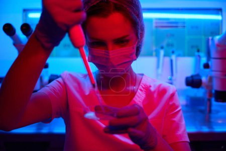 Foto de Asistente de laboratorio en una máscara protectora manipula el biomaterial, utiliza una taza de Petri y una pipeta especial - Imagen libre de derechos