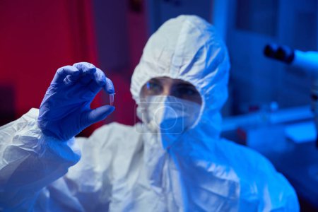 Foto de Asistente de laboratorio en uniforme protector sostiene tubo de ensayo con biomaterial en sus manos, en muestra de tubo de ensayo de líquido rojo - Imagen libre de derechos