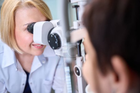 Téléchargez les photos : Optométriste féminine caucasienne expérimentée regardant à travers le microscope de lampe à fente aux yeux du patient - en image libre de droit