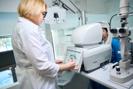 Photo for Vision specialist looking at adult patient eye image on autorefractor touch screen - Royalty Free Image