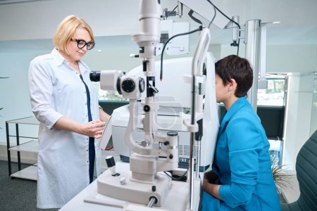 Téléchargez les photos : Optométriste souriante effectuant une réfraction objective automatisée chez une cliente adulte pendant la consultation - en image libre de droit