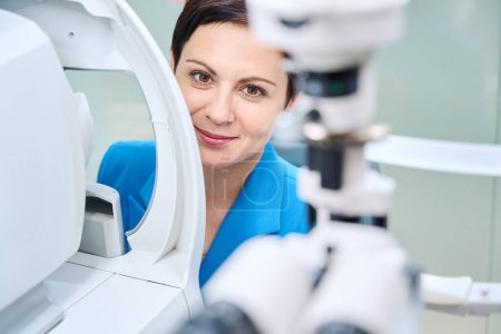 Foto de Retrato de una mujer adulta sonriente sentada frente al dispositivo oftálmico en la oficina de optometría - Imagen libre de derechos