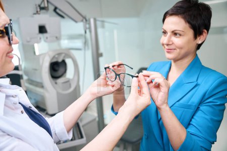 Téléchargez les photos : Ophtalmologiste amicale démontrant paire de lunettes à la cliente dans son bureau - en image libre de droit