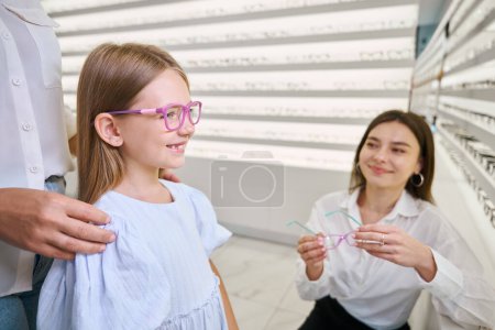 Téléchargez les photos : Joyeuse petite fille essayant une paire de lunettes en présence de son parent et d'une opticienne - en image libre de droit