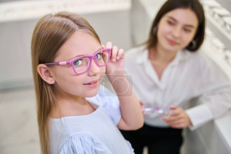 Téléchargez les photos : Enfant essayant une paire de lunettes devant la caméra en présence d'une opticienne - en image libre de droit