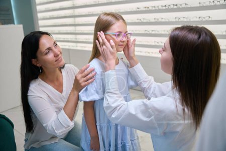Optical store consultant putting eyeglasses on female child face in presence of pleased mother