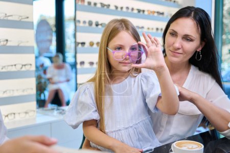 Téléchargez les photos : Petite fille assise avec mère à table dans un magasin d'optique regardant à travers une lentille teintée à la main en présence d'opticien - en image libre de droit