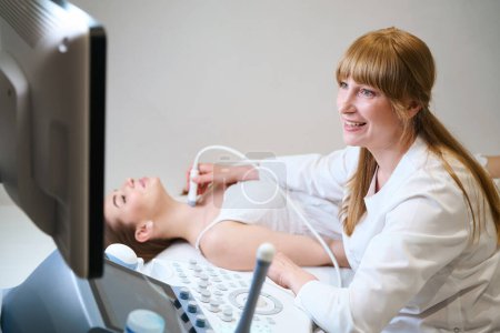 Hübscher Arzt führt Ultraschall der Schilddrüse zur werdenden Mutter, der Ultraschallraum ist hell und sauber