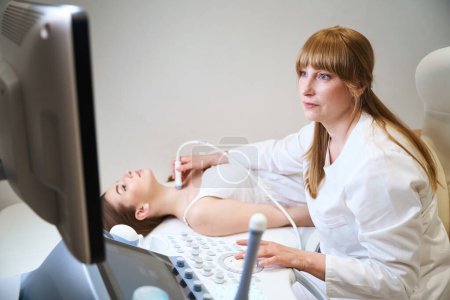 Foto de Rubia uzist trabaja en un dispositivo de ultrasonido moderno, ella hace un ultrasonido de la glándula tiroides para un paciente joven - Imagen libre de derechos