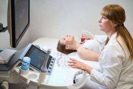 Téléchargez les photos : Blonde utilise un appareil ultrasonore moderne dans son travail, elle fait une échographie de la glande thyroïde pour un jeune patient - en image libre de droit