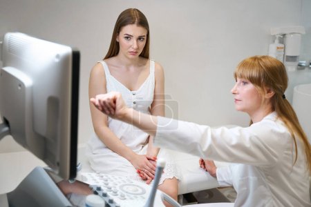 Foto de Mujer médico consulta a mujer joven en la sala de ultrasonido, el médico de ultrasonido señala el monitor de la máquina de ultrasonido - Imagen libre de derechos