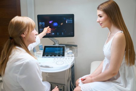 Téléchargez les photos : Femme blonde consulte une jeune femme dans une salle d'échographie, le médecin pointe vers le moniteur de la machine à échographie - en image libre de droit