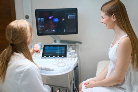 Foto de Médico femenino consulta joven feliz en la sala de ultrasonido, el médico señala el monitor de la máquina de ultrasonido - Imagen libre de derechos