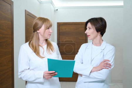 Téléchargez les photos : Les femmes en uniforme médical communiquent dans le couloir de l'hôpital, elles ont un dossier bleu avec des documents - en image libre de droit