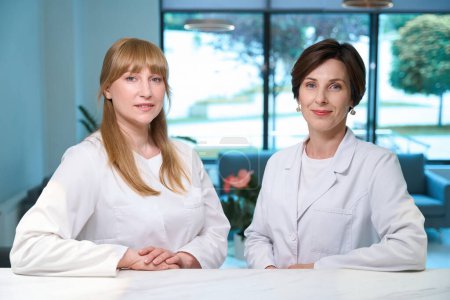 Téléchargez les photos : Deux belles femmes en uniforme médical debout à la réception, dans le foyer de l'hôpital intérieur respectueux de l'environnement - en image libre de droit