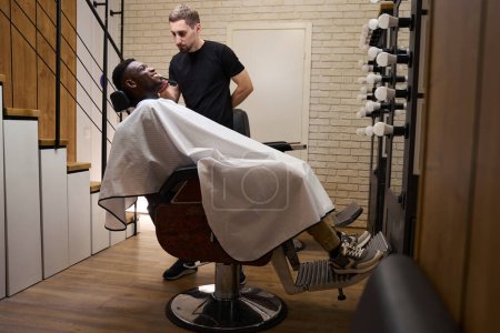 Photo for Barber shaves an African American client with an electric razor as he works in front of an illuminated mirror - Royalty Free Image