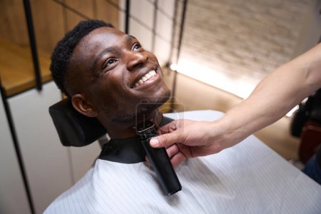 Foto de Sonriente cliente afroamericano se sienta cómodamente en una silla de barbero, un barbero lo afeita con una navaja eléctrica especial - Imagen libre de derechos