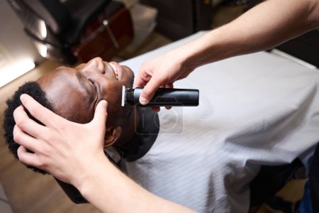 Photo for African American male sits in a barbershop in a barber chair, the barber shaves him with a special razor - Royalty Free Image