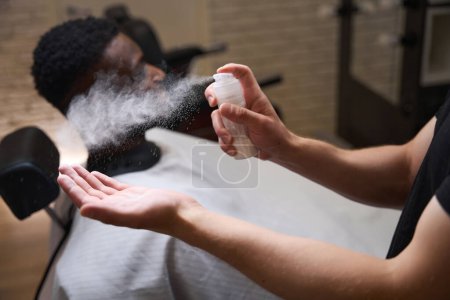 Téléchargez les photos : Afro-Américain assis dans un salon de coiffure dans un fauteuil de coiffeur, le coiffeur utilise un spray spécial dans son travail - en image libre de droit