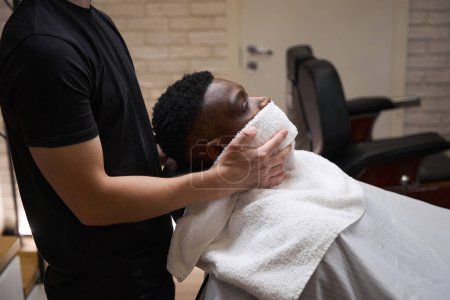 Foto de El chico afroamericano se sienta en una barbería en una silla de barbero, el barbero se puso una toalla caliente en la cara - Imagen libre de derechos
