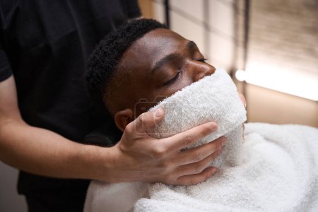 Photo for Barber put a warm towel on the face of the African American guy, the client sits with his eyes closed - Royalty Free Image