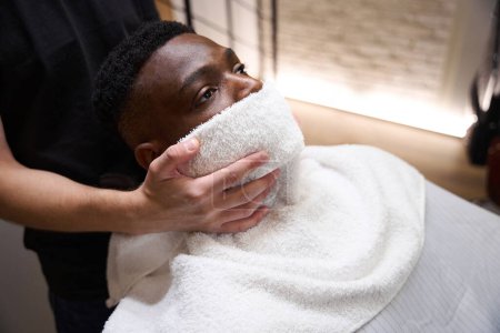 Foto de Un peluquero masculino puso una toalla caliente en la cara de un afroamericano, un cliente con una capa protectora - Imagen libre de derechos