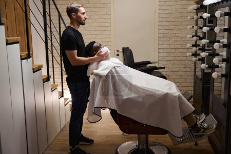 Photo for Male barber put a warm towel on the face of an African American guy, a client in a protective cape - Royalty Free Image