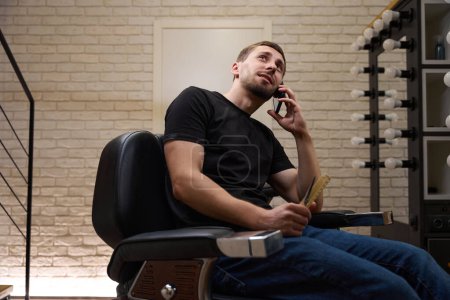 Foto de El tipo está hablando por teléfono, sentado en una silla de peluquero, está en su lugar de trabajo en una barbería - Imagen libre de derechos