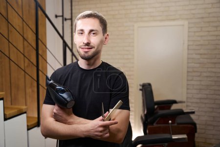 Photo for Barber holds a hair dryer, scissors and a comb in his hands, this is his working tool - Royalty Free Image