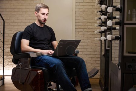 Foto de El tipo se comunica en línea en un ordenador portátil, sentado en una silla de peluquería, que está en su lugar de trabajo en una barbería - Imagen libre de derechos