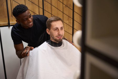 Foto de Joven en un velo de protección se sienta en una silla de barbero, un peluquero afroamericano aconseja a un cliente - Imagen libre de derechos