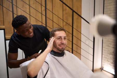 Téléchargez les photos : Beau homme au voile protecteur assis sur une chaise de coiffeur, il communique avec un coiffeur afro-américain - en image libre de droit