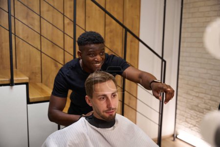 Photo for African American hairdresser selects a hairstyle for a client with a beard, the room has a minimalist interior - Royalty Free Image