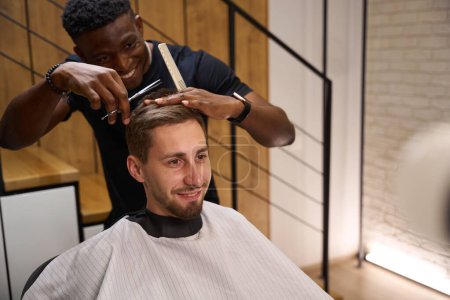 Foto de Joven peluquero afroamericano corta una tijera clientes, la peluquería tiene un interior minimalista - Imagen libre de derechos