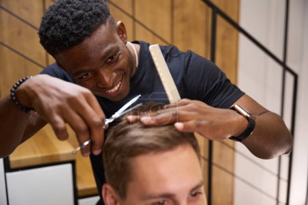 Photo for Cheerful African American hairdresser makes a haircut with scissors for a young man, a minimalist interior in a barbershop - Royalty Free Image