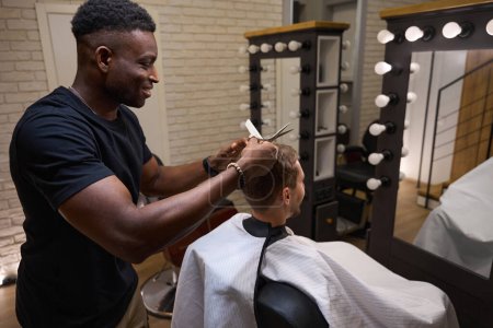 Photo for African American hairdresser gives a haircut to a young man, the master uses scissors and a comb - Royalty Free Image