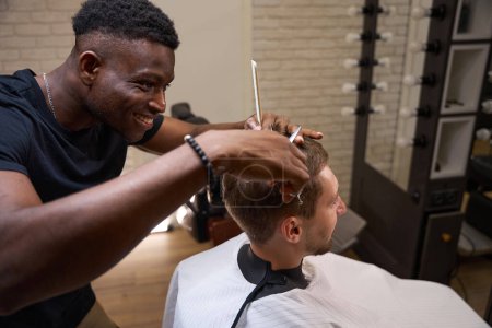 Photo for Smiling African American hairdresser gives a haircut to a young man, the master uses scissors and a comb - Royalty Free Image