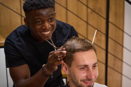 Photo for Friendly African American guy makes a haircut for a client, the master uses scissors and a comb - Royalty Free Image