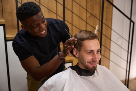 Foto de Amistoso maestro afroamericano hace un corte de pelo para un cliente, un tipo utiliza tijeras y un peine - Imagen libre de derechos