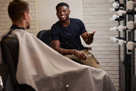 Photo for African American man and a caucasian male communicate while sitting in barber chairs, barber holding hairdressing tools - Royalty Free Image
