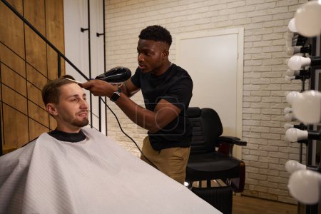 Photo for Young African American hairdresser does hair styling for client, in his work the guy uses hair dryer and a comb - Royalty Free Image