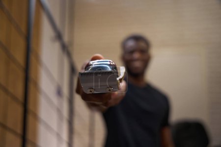 Foto de Sonriente peluquero afroamericano sostiene una navaja eléctrica especial en sus manos, el tipo está en el lugar de trabajo en la barbería - Imagen libre de derechos